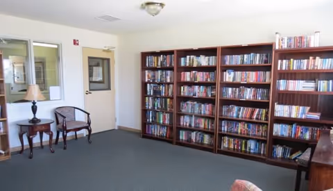 Community reading room with a wall of bookshelves, two chairs, a side table with a lamp, and a door.