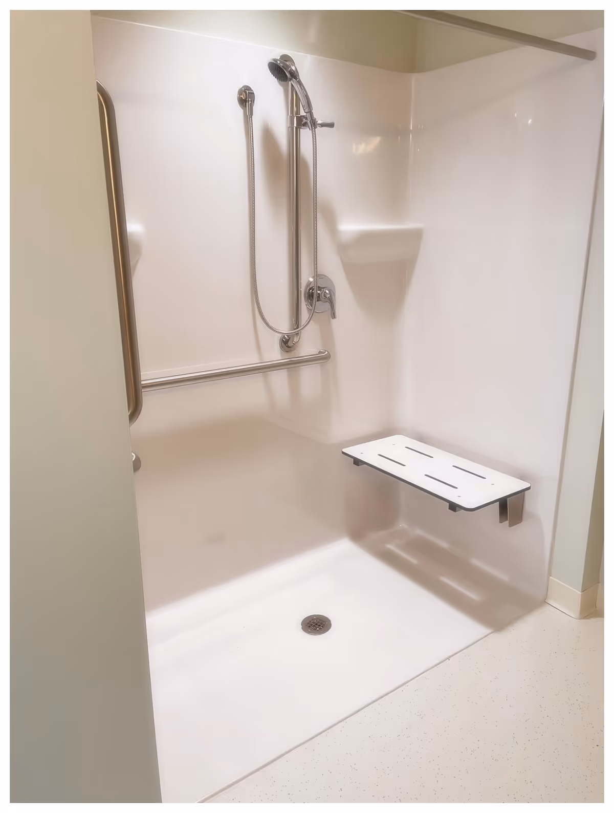 A clean, accessible shower area with a handheld showerhead, a horizontal grab bar, a built-in corner shelf, and a fold-down shower seat mounted on the wall.