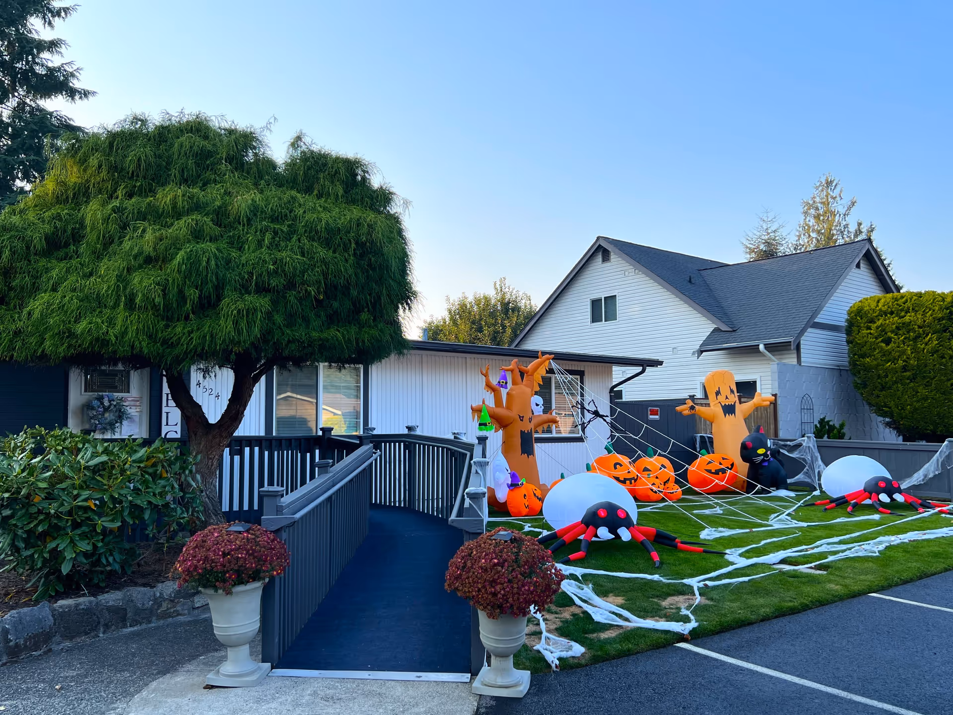 Exterior view of a senior care home decorated for Halloween with inflatable pumpkins, spiders, a tree, and a ghost on a green lawn. The building has a ramp leading to the entrance, and there are potted plants on either side of the ramp.