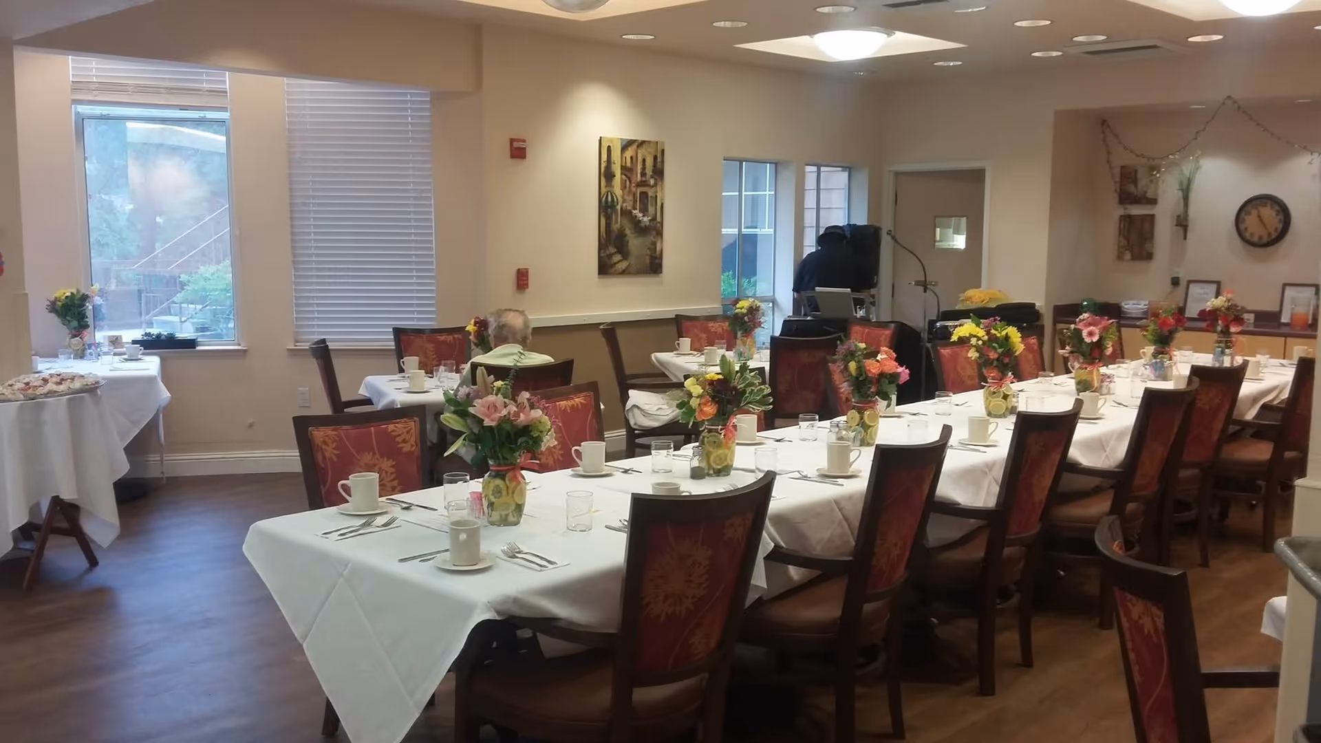 A dining room in an assisted living facility with long tables covered in white tablecloths, set with cups, glasses, and silverware. Each table has multiple floral centerpieces. There are several chairs with red and gold patterned upholstery around the tables. A few people are seated, and there are windows letting in natural light. The room has light-colored walls with some decorations and a clock on the wall.