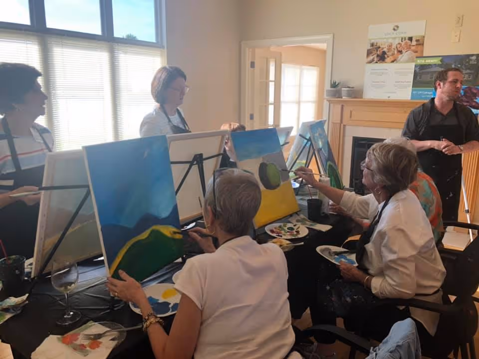 A group of elderly people seated around a table in a well-lit room, engaged in a painting activity with canvases on easels. They are painting landscapes with blue skies and green hills. A man stands nearby, possibly instructing or assisting the group. The room has large windows with blinds and a fireplace with informational posters on the mantel.