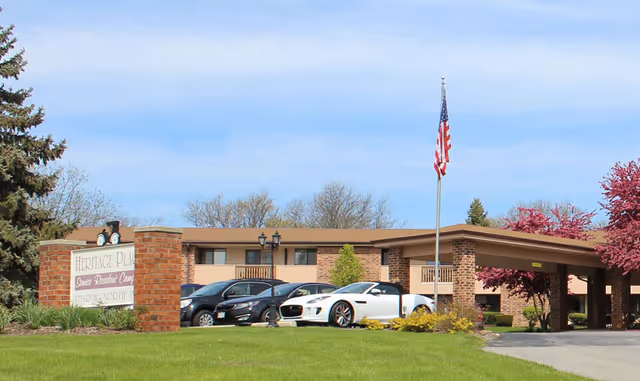 Front exterior of Ruby Commons senior living building with entrance canopy, parked cars, a flagpole, sign, and landscaped lawn.