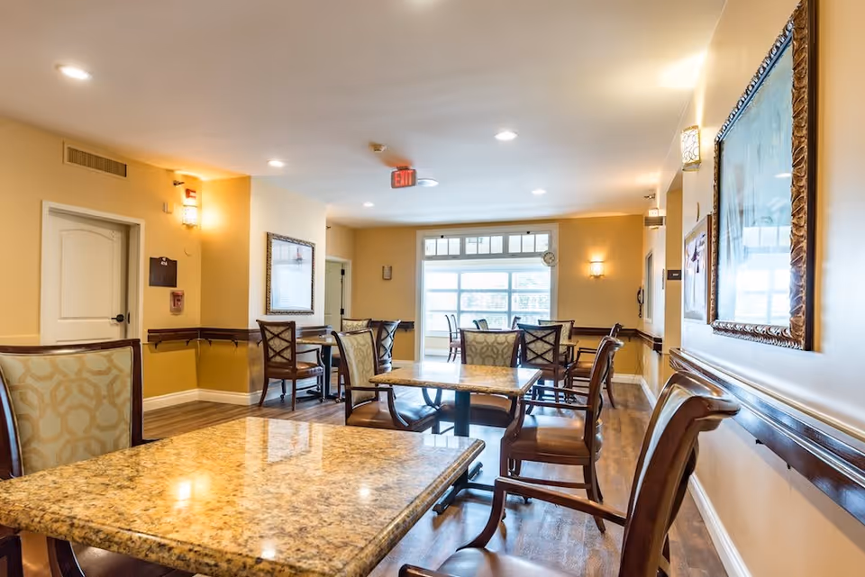 A well-lit dining area in a senior living facility with several tables and chairs arranged neatly. The room has warm beige walls, wooden flooring, framed artwork on the walls, and large windows letting in natural light. There are handrails along the walls and ceiling lights providing additional illumination.
