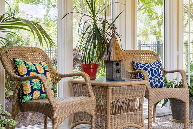 Sunroom with two wicker chairs and a wicker table holding a decorative birdhouse and potted plants in front of large windows.