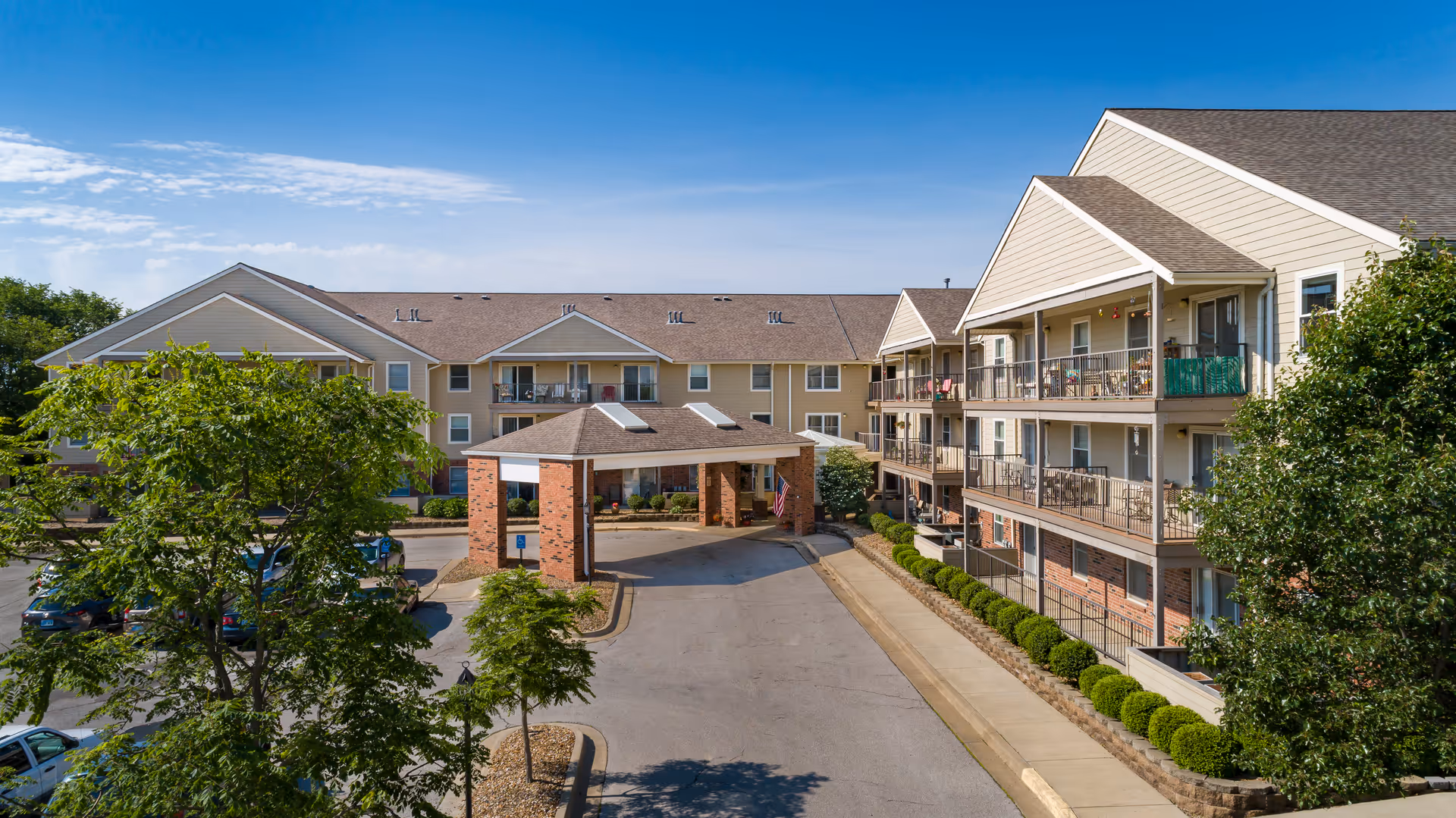 Front exterior of a multi-story senior living building with a covered drive-up entrance, balconies, landscaping, and parked cars.