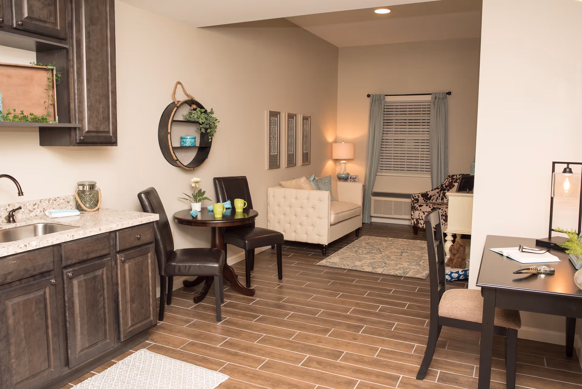 Interior view of a senior living facility room featuring a small kitchenette with dark wood cabinets and a granite countertop, a round wooden table with two dark leather chairs, a beige tufted loveseat with decorative pillows, a patterned armchair, a desk with a chair and lamp, and a window with light blue curtains and blinds. The floor is covered with wood-look tiles and there is a small rug in front of the loveseat.