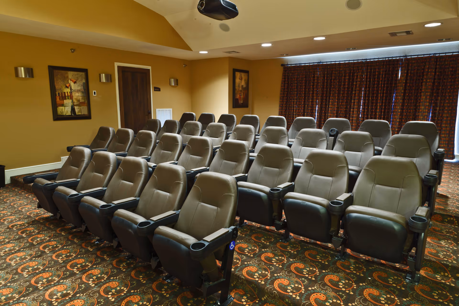 A small theater room with multiple rows of comfortable, padded reclining seats arranged in a tiered layout. The room has patterned carpet, beige walls with framed artwork, and a ceiling-mounted projector. Heavy curtains cover the windows at the back of the room.