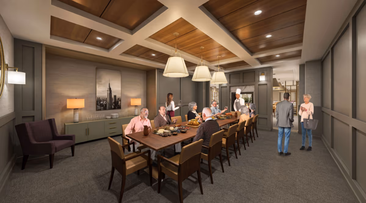 A dining room in a senior living facility with residents seated around a long table and staff standing nearby.