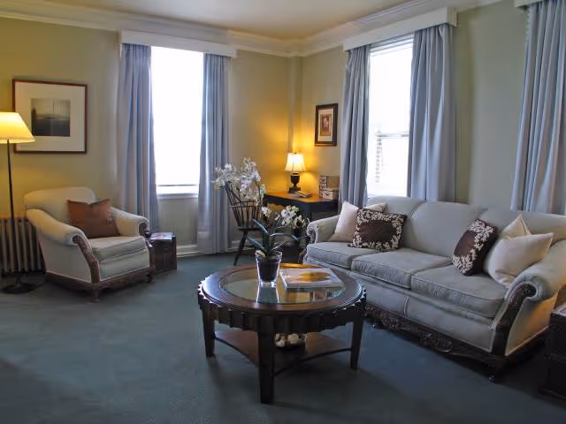 Sunlit living room with a sofa, armchair, round coffee table and windows with curtains.