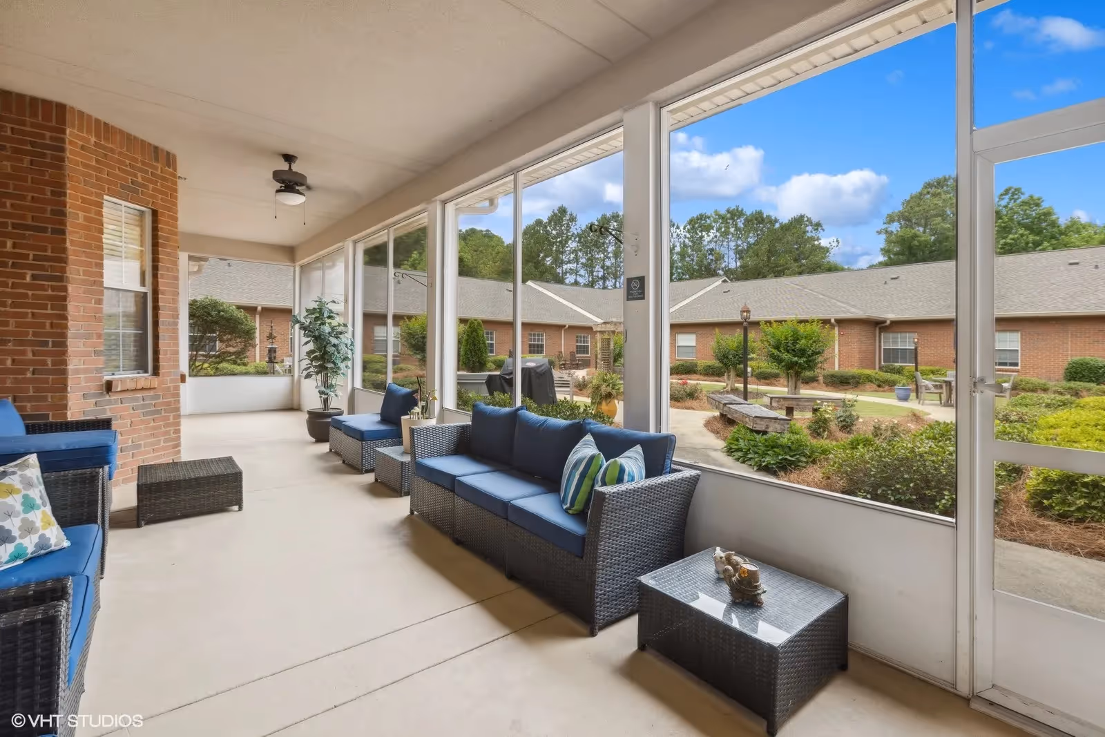 A screened-in porch area with blue cushioned wicker furniture including a sofa, chairs, and a coffee table. The porch overlooks a landscaped courtyard with shrubs, trees, and a brick building in the background under a partly cloudy sky.