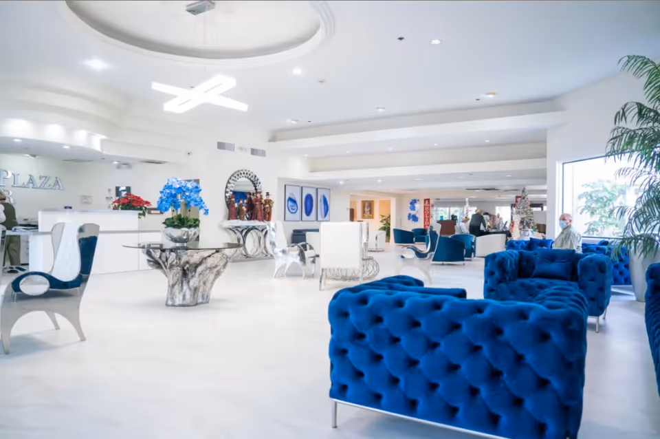 Bright and spacious senior living facility lobby with modern blue velvet tufted sofas, white chairs, a glass table with a decorative base, potted plants, and a reception desk in the background with the word 'PLAZA' visible on the wall.