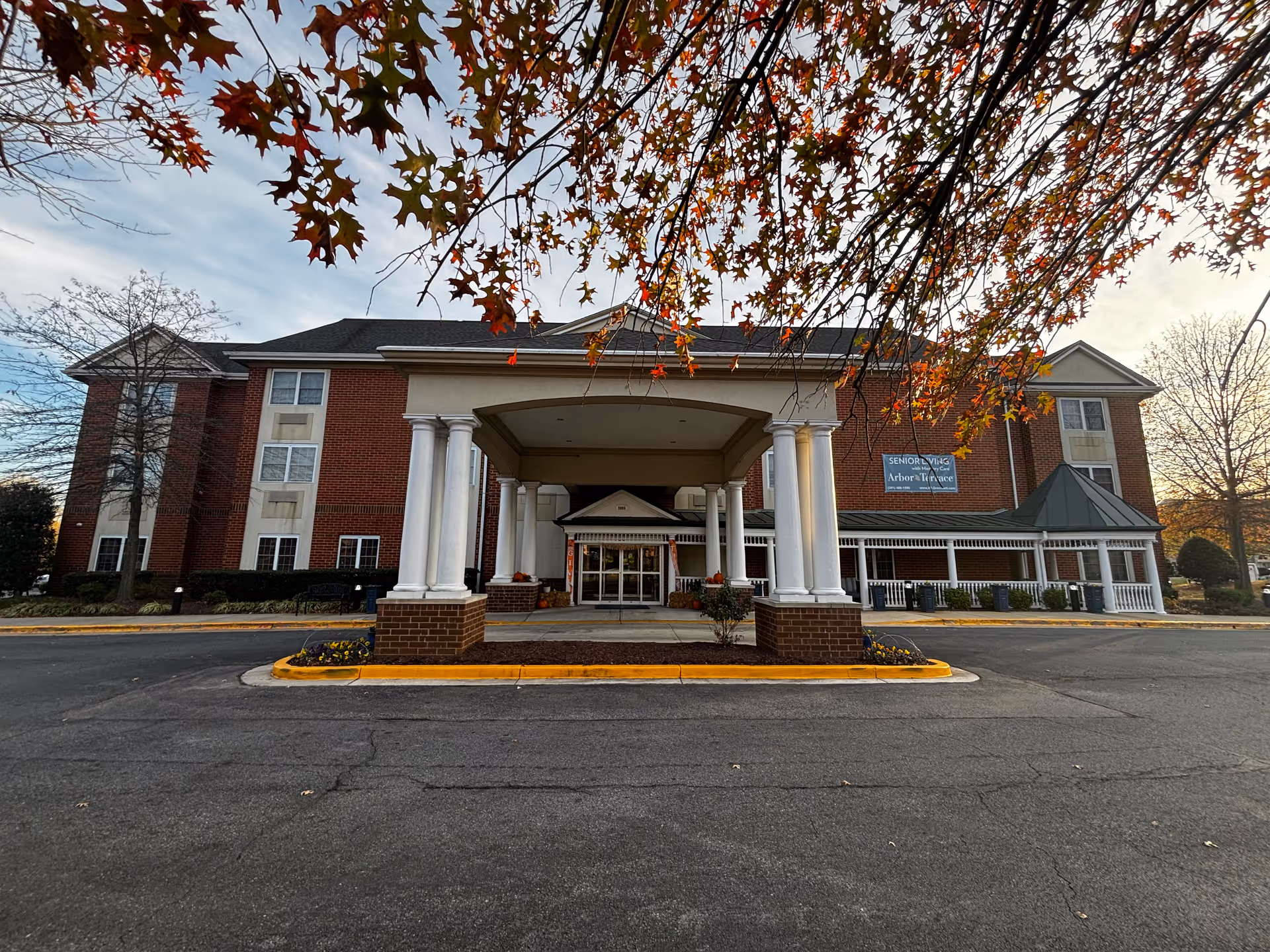 Front exterior view of a senior living facility building with a covered entrance supported by white columns. The building is made of red brick with beige accents and has multiple windows. There are autumn leaves hanging from a tree branch in the foreground and a paved driveway leading up to the entrance.