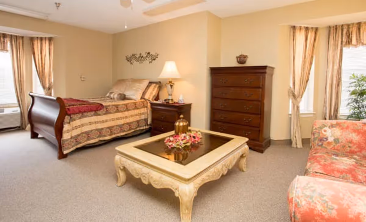 A cozy bedroom with a wooden sleigh bed covered in a patterned bedspread, a wooden nightstand with a lamp, a tall wooden dresser, a decorative coffee table with flowers, and a floral upholstered sofa. The room has beige walls, carpeted floor, and windows with curtains allowing natural light.