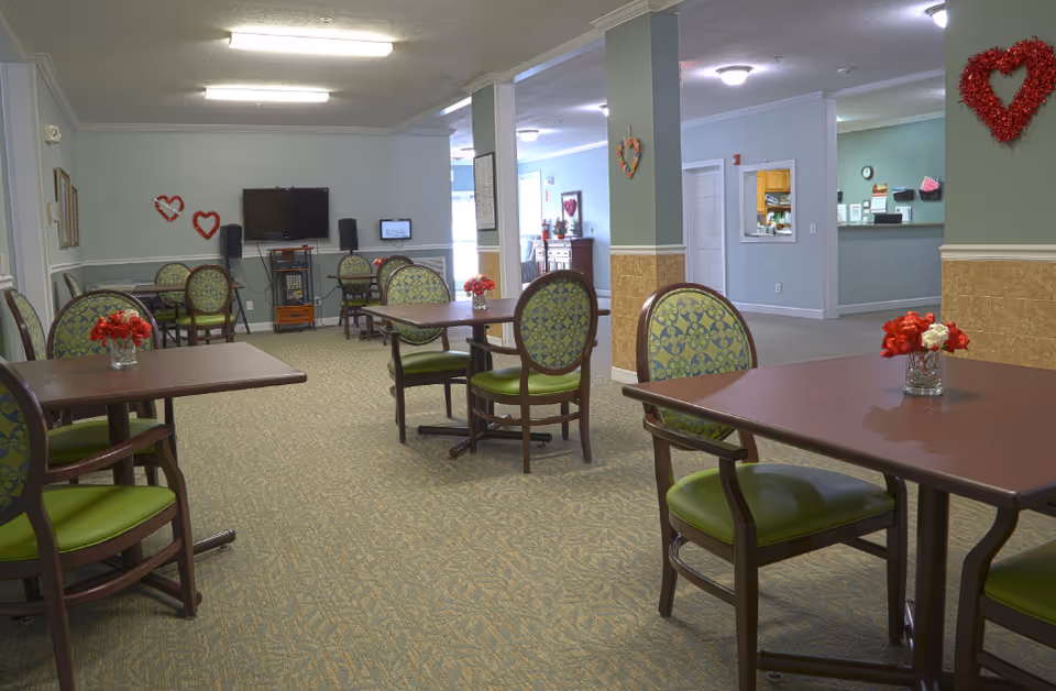 A senior living facility common area with several tables and chairs arranged for dining or socializing. The chairs have green patterned upholstery and the tables have small vases with red flowers. The walls are decorated with red heart-shaped ornaments. A television and speakers are mounted on the far wall, and there is a reception area visible in the background.