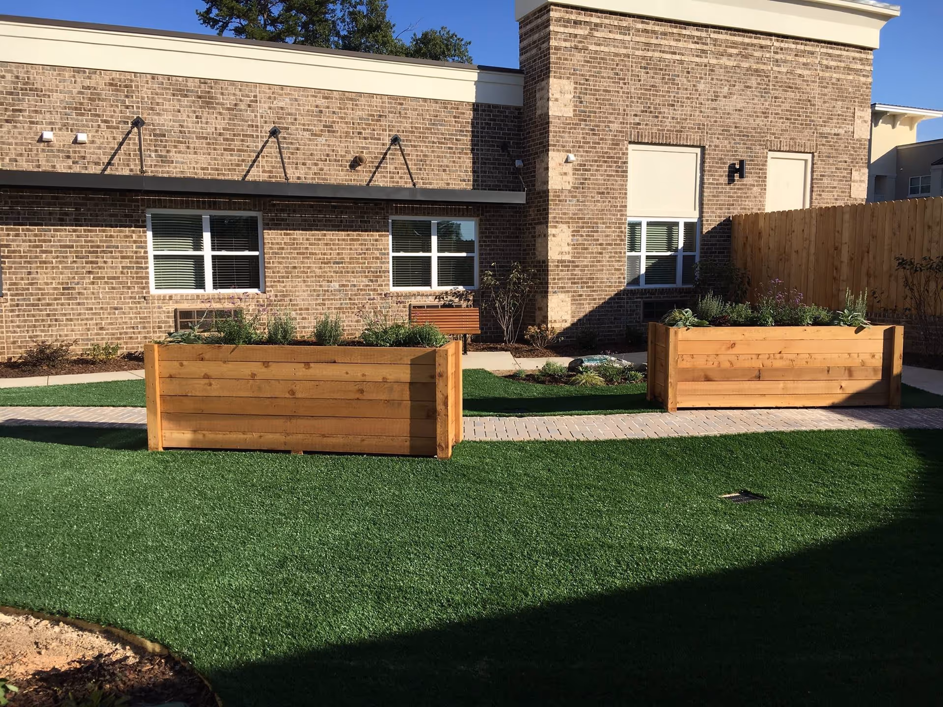 Outdoor garden area with two large wooden planter boxes filled with plants, artificial green grass, a paved walkway, and a brick building with windows and a wooden fence in the background.