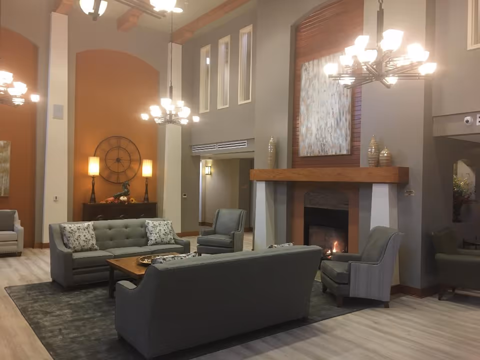 Spacious furnished lounge with sofas and armchairs arranged around a coffee table and a lit fireplace beneath chandeliers.