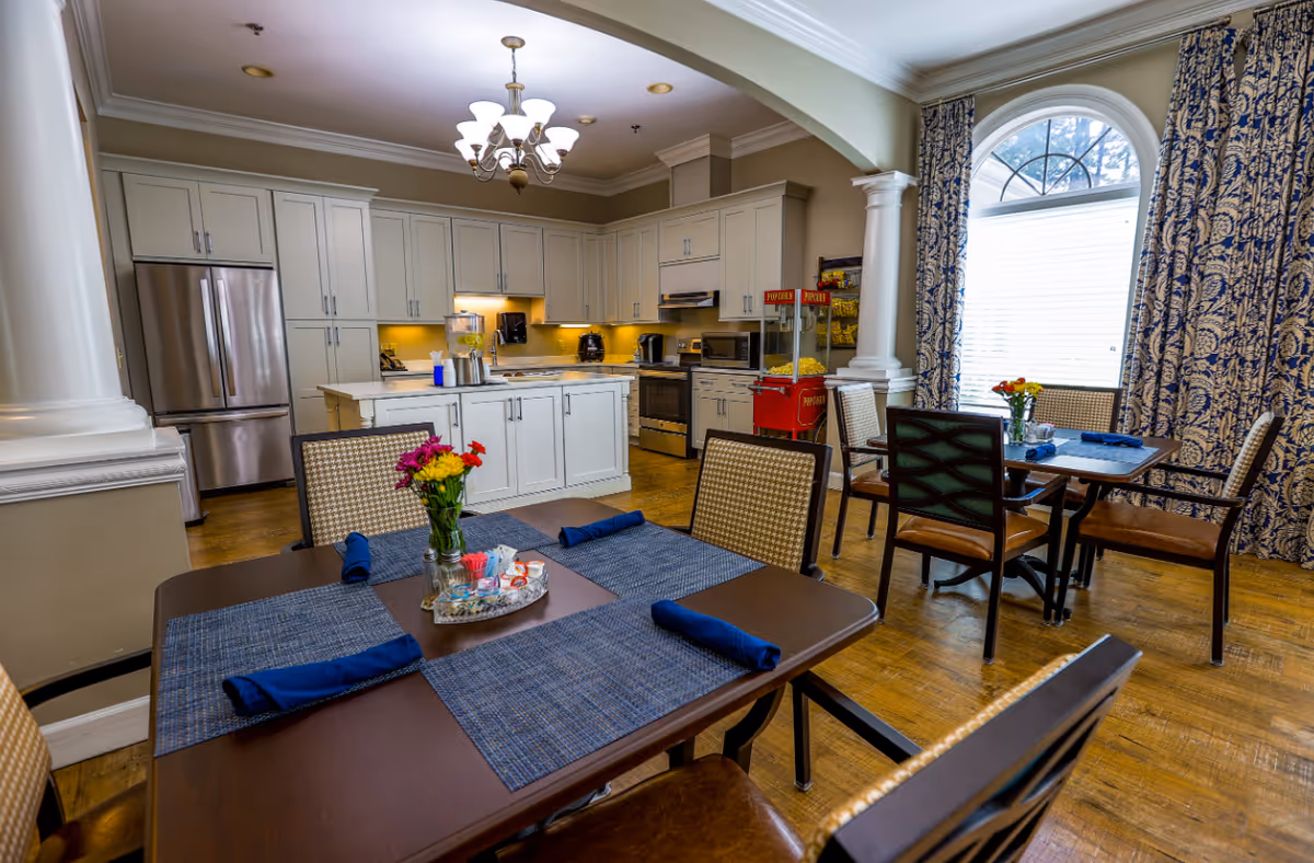 A bright and inviting dining area with a kitchen in the background. The kitchen features white cabinetry, stainless steel appliances, and a countertop with a beverage dispenser. The dining area has tables set with blue placemats, rolled blue napkins, and small flower arrangements. Large windows with patterned blue and white curtains allow natural light to fill the space. A popcorn machine is visible near the kitchen area.