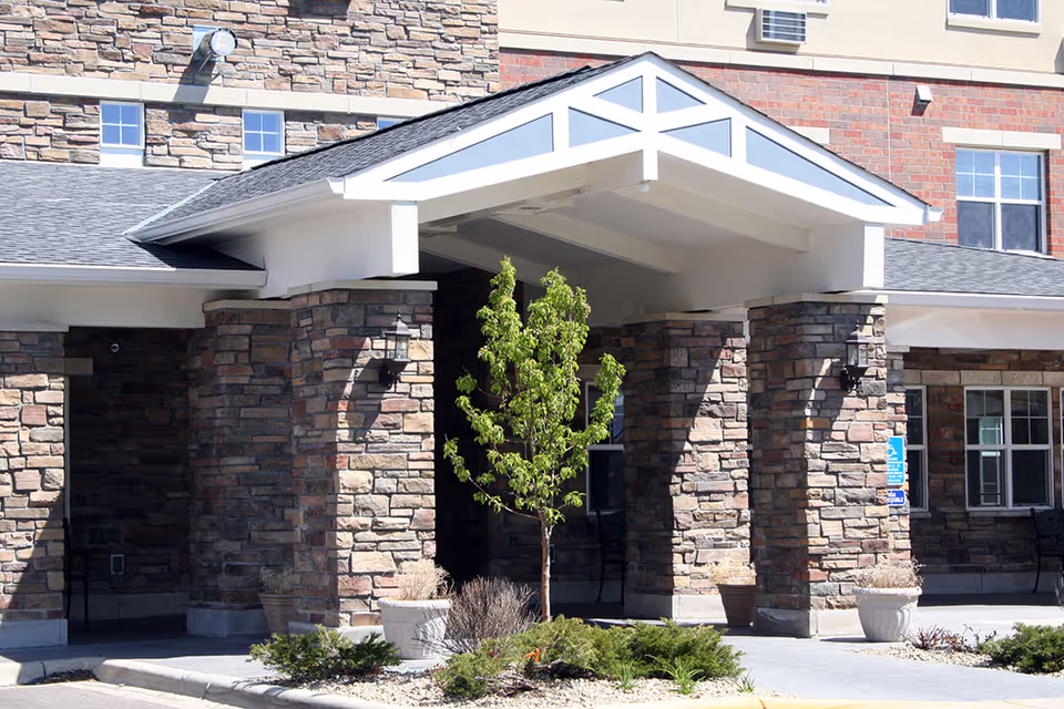 Entrance of a senior living facility with stone pillars, a covered porch, a small tree, and potted plants in front of the building.