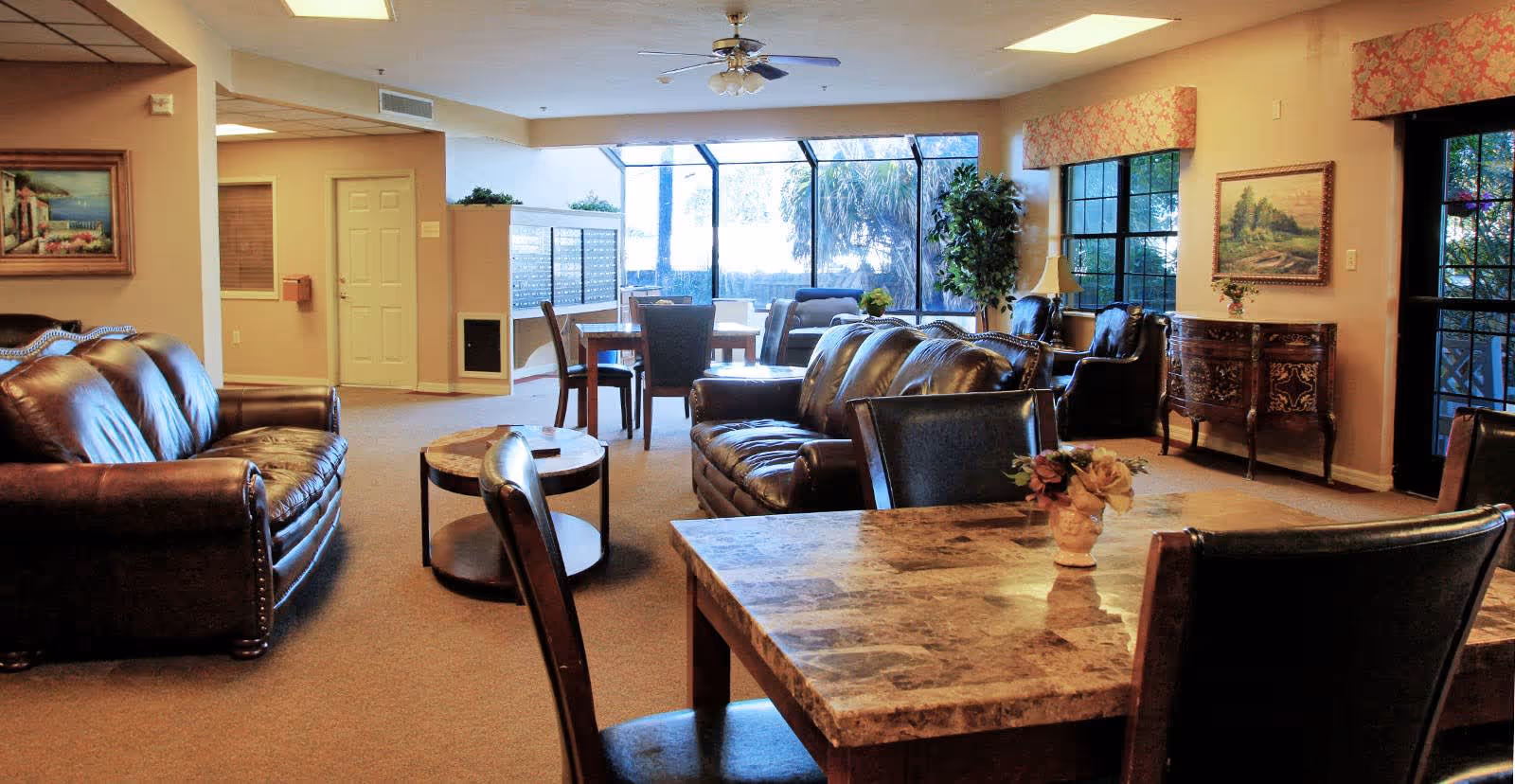 Bright communal living room with leather sofas, dining tables, large windows, and decorative plants and artwork.