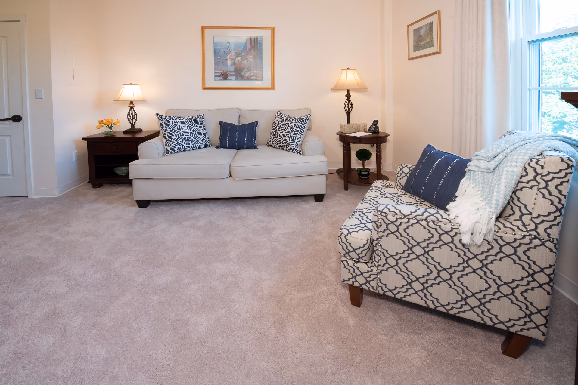 A cozy living room with a beige sofa adorned with three decorative pillows, two side tables each with a lamp, a patterned armchair with a blue pillow and a light blue throw blanket, a window with white curtains, and framed artwork on the walls.