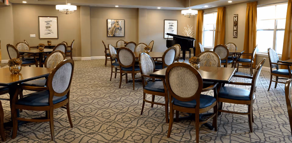 A spacious dining room with multiple wooden tables and cushioned chairs arranged neatly on a patterned carpet. The room is well-lit with natural light coming through large windows with golden curtains. There are framed artworks on the walls and a black grand piano in the corner. A decorative sign with the word 'HOME' is mounted on the wall near the windows.