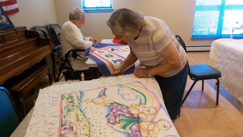 Two elderly women engaged in quilting activities in a well-lit room. One woman is standing and working on a quilt with teddy bear and rainbow designs, while the other woman is seated at a table working on a different quilt. The room has wooden flooring, a piano against the wall, and windows with blue blinds.