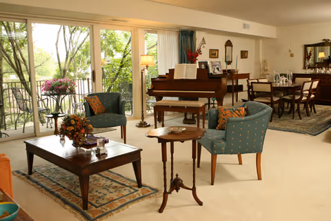 A spacious and well-lit living room with large windows showing green trees outside. The room features a wooden coffee table with a floral arrangement, two upholstered armchairs with patterned cushions, a small round side table, and a grand piano with sheet music. In the background, there is a dining area with a wooden dining table and chairs, a sideboard with decorative items, and a large mirror on the wall.