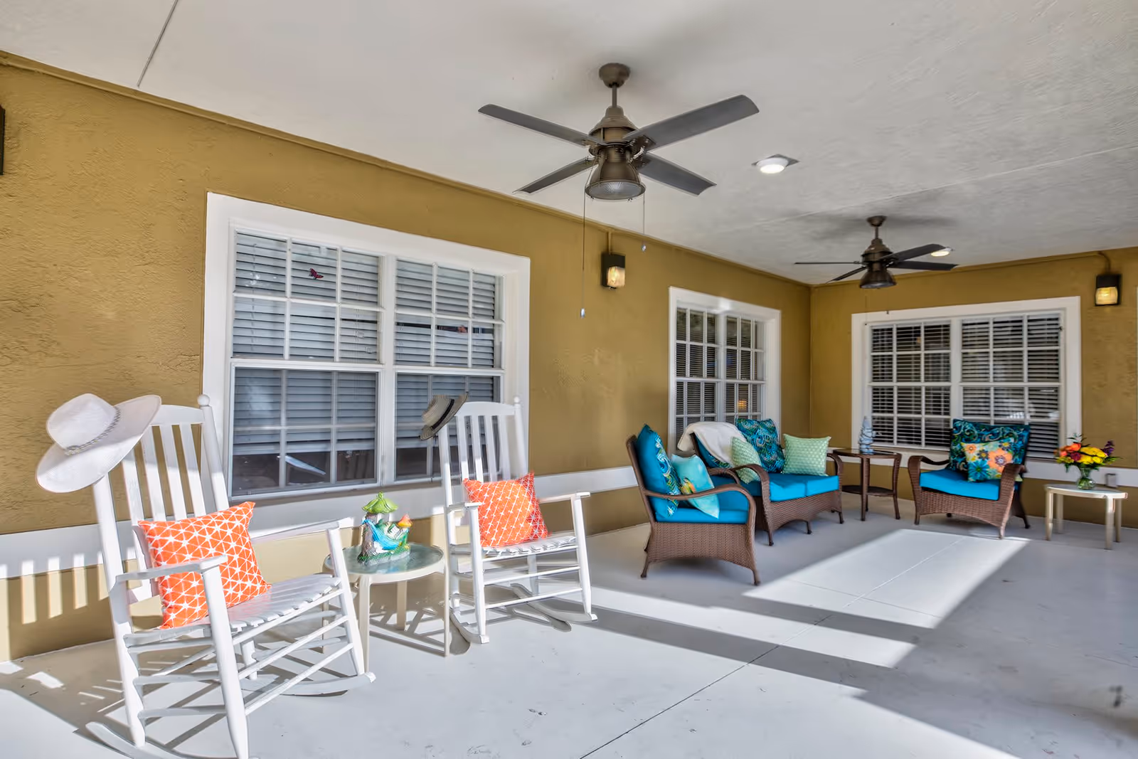 Covered outdoor patio area with white rocking chairs adorned with orange pillows and hats, a small glass-top table with a decorative item, wicker armchairs and loveseat with blue cushions and colorful pillows, a small side table with a flower vase, ceiling fans, and windows with white blinds on a beige wall.