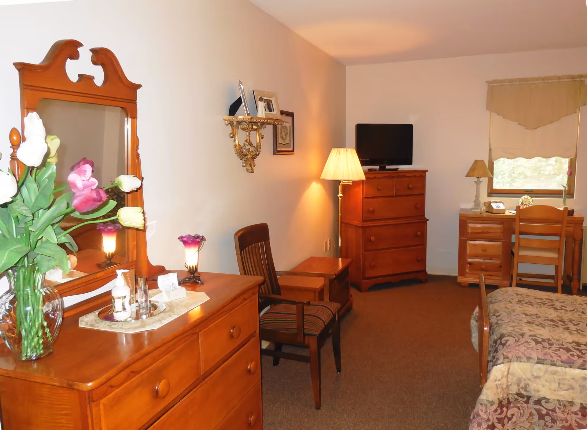 A cozy bedroom with wooden furniture including a dresser with a mirror, a chair, a chest of drawers with a TV on top, and a desk with a chair near a window. The room is softly lit by a floor lamp and a table lamp, and there is a vase with colorful flowers on the dresser.