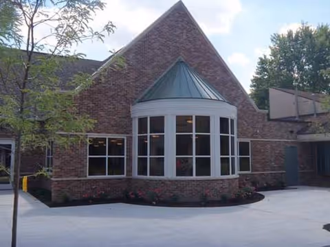 Brick senior living facility with a curved bay window, triangular copper-green roof, and surrounding landscaping.