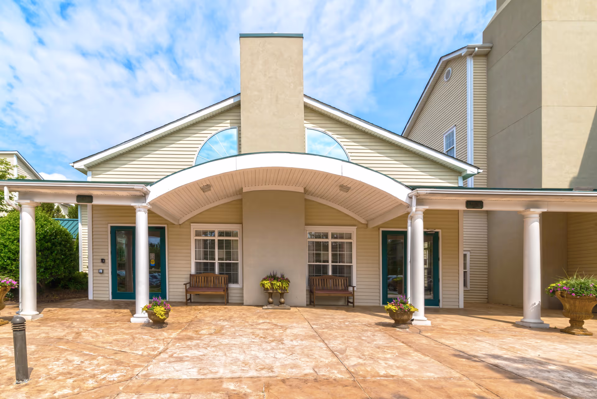 Front entrance of a senior living building with an arched canopy, columns, two benches, and potted planters.