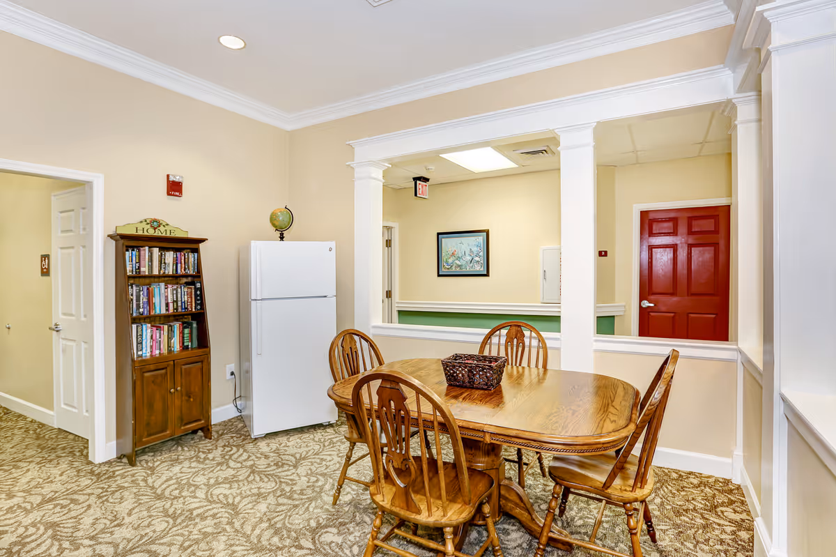 A cozy dining area with a wooden table and four matching chairs on a patterned carpet. There is a white refrigerator next to a wooden bookshelf filled with books and topped with a decorative globe and a sign that says 'HOME'. The walls are painted beige, and there is a red door and a framed picture visible through an open partition with white columns.