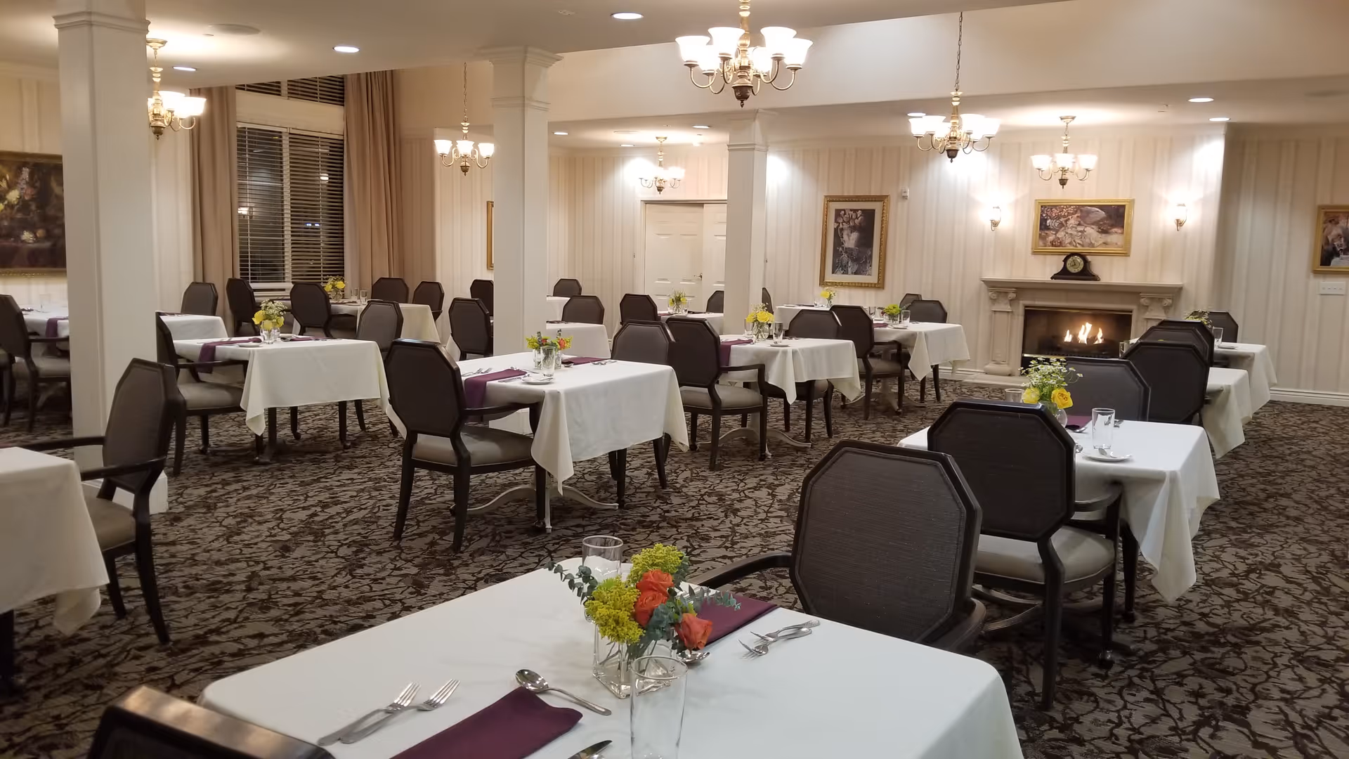 A dining room with multiple tables covered in white tablecloths and set with silverware, glasses, and purple napkins. Each table has a small floral centerpiece. The room features carpeted floors, beige striped wallpaper, framed artwork on the walls, and several chandeliers providing warm lighting. A fireplace with a clock on the mantel is visible in the background.