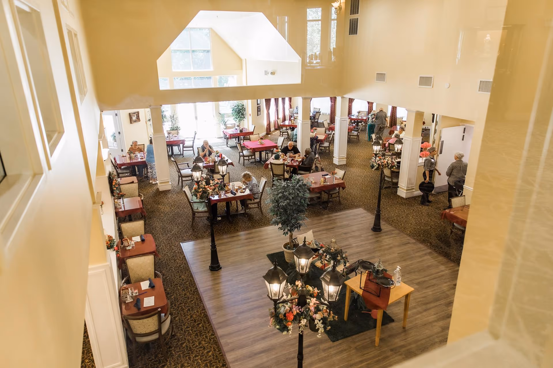 A spacious dining room in a senior living facility with multiple tables covered in red tablecloths. Several elderly residents are seated at the tables eating and socializing. The room has high ceilings, large windows letting in natural light, and decorative streetlamp-style lights with floral arrangements in the center. Staff members are seen walking and assisting residents.