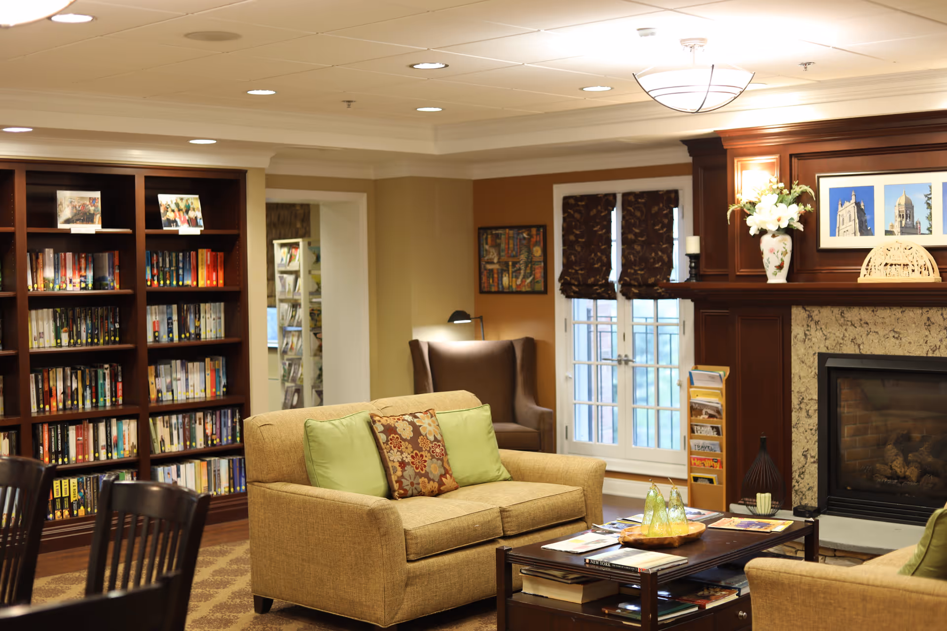 A cozy living room area in a senior living facility featuring beige sofas with green and floral cushions, a dark wooden coffee table with magazines, a fireplace with a wooden mantel decorated with flowers and framed pictures, a bookshelf filled with books, and a comfortable armchair near a window with patterned curtains.