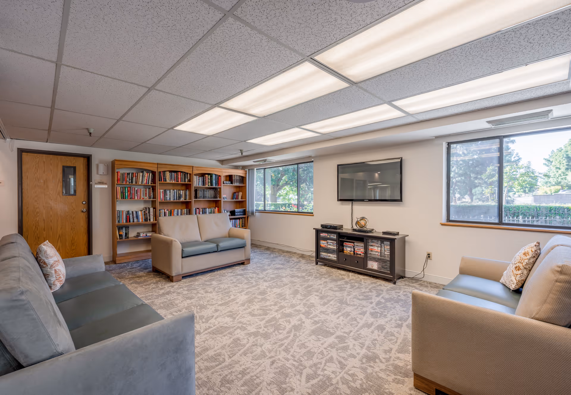 A cozy living room area in a senior living facility featuring two large windows with views of greenery outside, two beige sofas with patterned pillows, a wooden bookshelf filled with books, a flat-screen TV mounted on the wall above a black media console with DVDs and decorative items, and a carpeted floor with a subtle pattern.