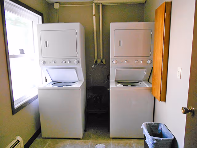 Laundry room with two stacked washer and dryer units side by side, a window on the left wall, a wooden cabinet on the right wall, and a trash bin on the floor near the door.