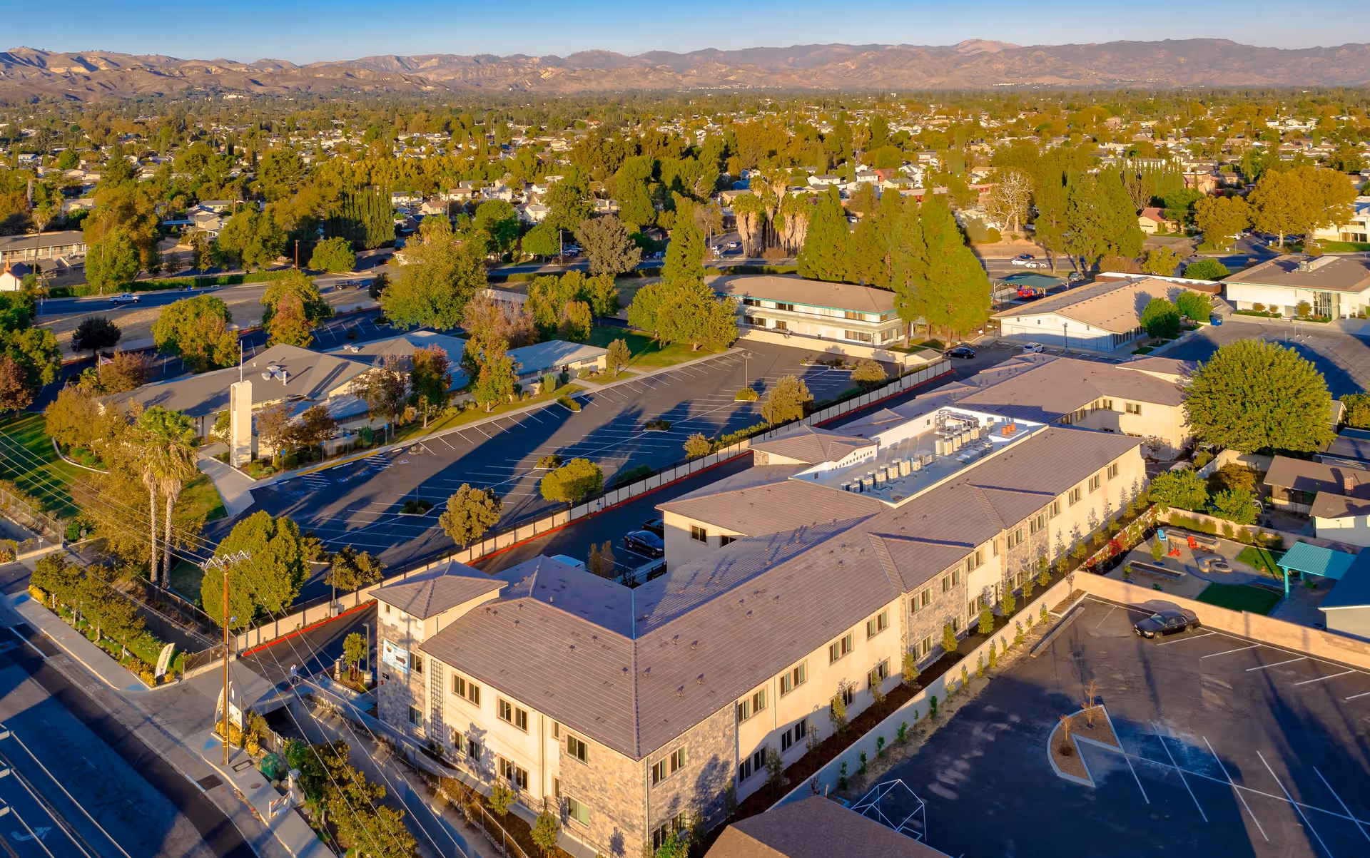 Aerial view of Vista At Simi Valley senior living facility showing multiple buildings with beige roofs, surrounding parking lots, green trees, and a playground area, with mountains visible in the background under a clear sky.