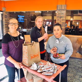 Three women standing behind a cart filled with Sonic fast food bags inside a facility with brick pillars and a TV in the background. One woman is smiling and wearing glasses, another is holding a wrapped item, and the third is holding a phone and some food wrappers.