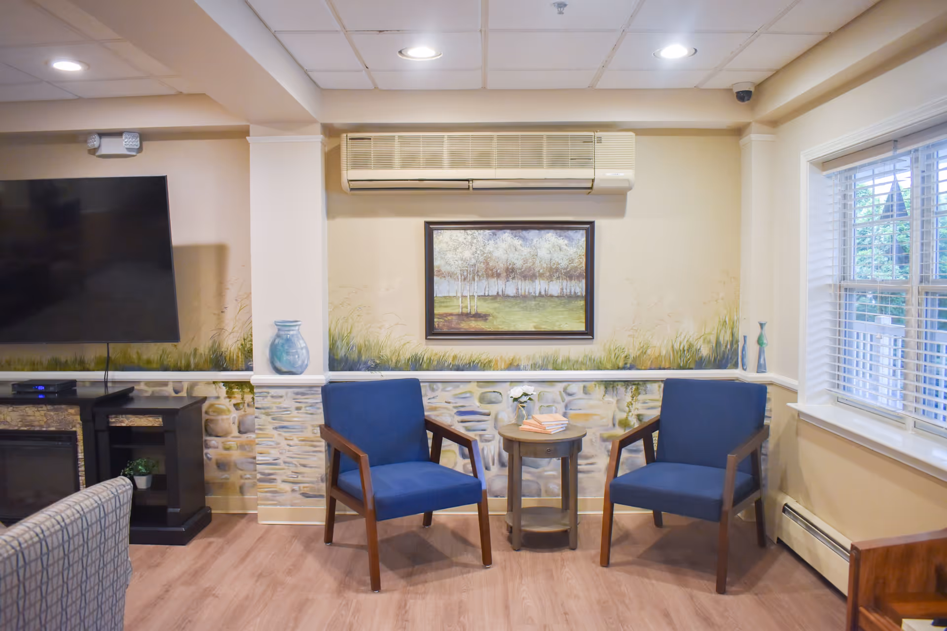 A cozy common room with two blue armchairs flanking a small round table beneath a framed painting and wall air conditioner, a TV to the left and a window to the right.