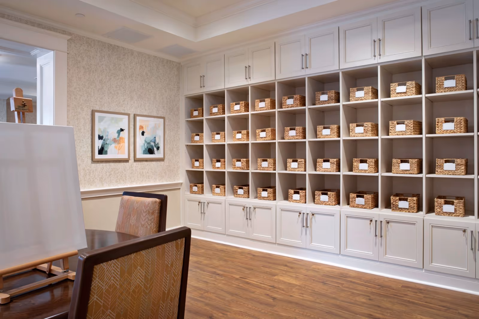 Light-filled interior activity room with built-in cubby shelves of labeled wicker baskets, a table with chairs and an easel.