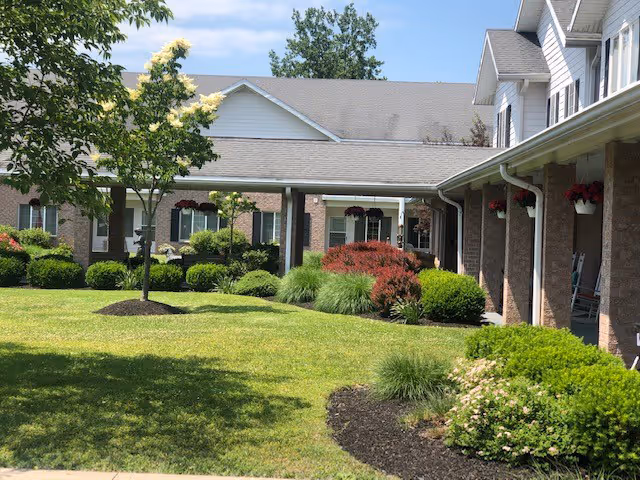 Outdoor view of a senior living facility with well-maintained green lawn, various bushes, small trees, and a covered walkway connecting brick buildings with white trim and hanging flower pots.