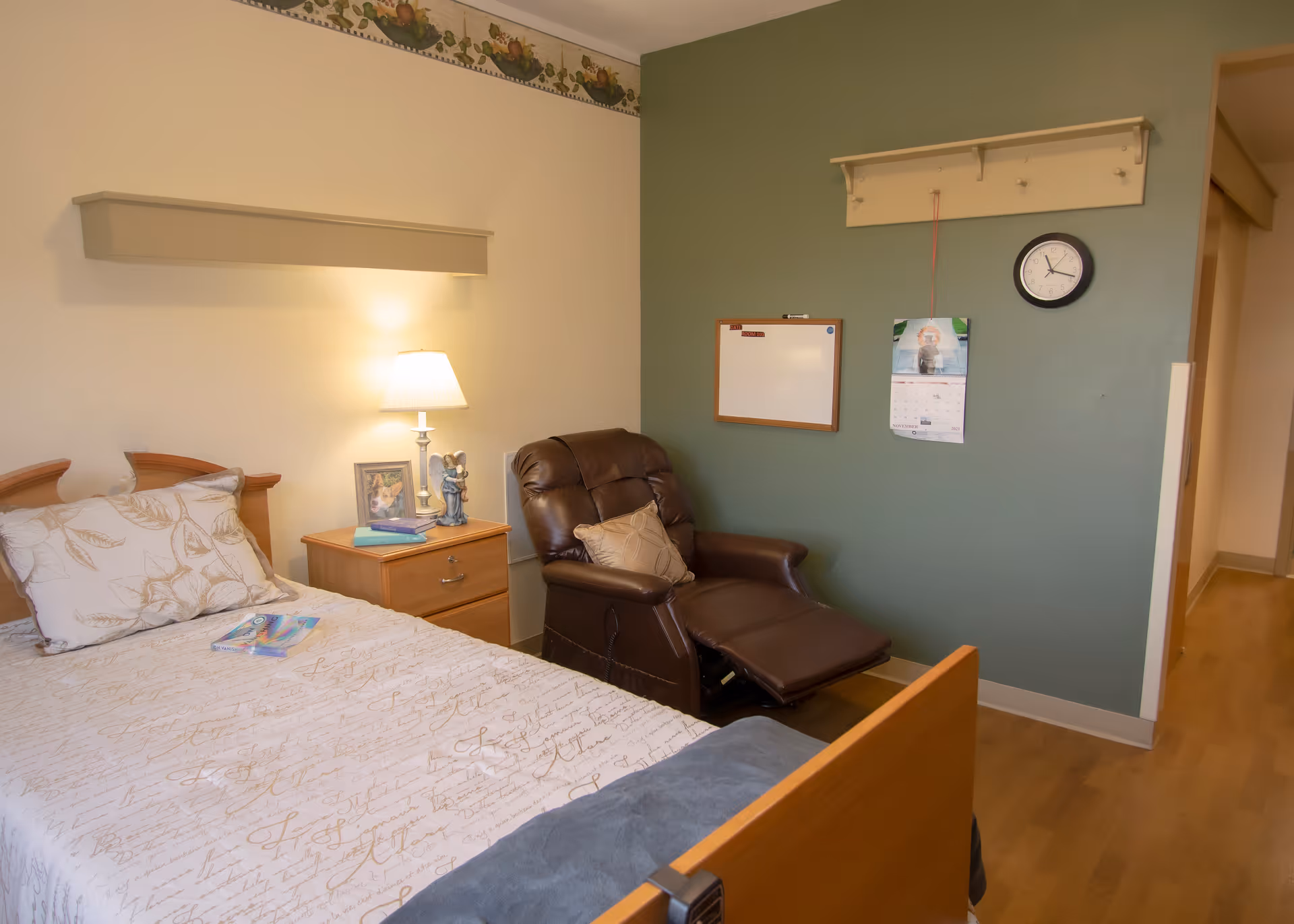 A cozy bedroom in a senior living facility featuring a single bed with patterned bedding, a wooden nightstand with a lamp, books, and a framed photo. Next to the nightstand is a brown recliner chair with a cushion. The walls are painted in two tones, with a green accent wall holding a whiteboard, a calendar, a clock, and a coat rack. The floor is wooden.