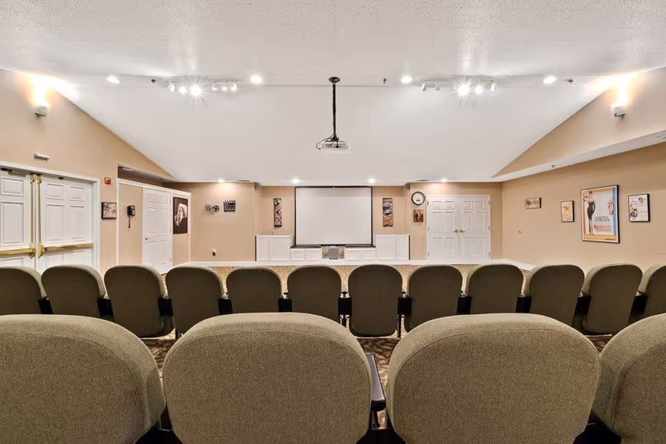 A small auditorium or screening room with rows of cushioned chairs facing a large white projection screen mounted on a beige wall. The room has beige walls, carpeted floor, ceiling lights, and framed pictures on the walls. There are double doors on either side of the screen and a projector hanging from the ceiling.
