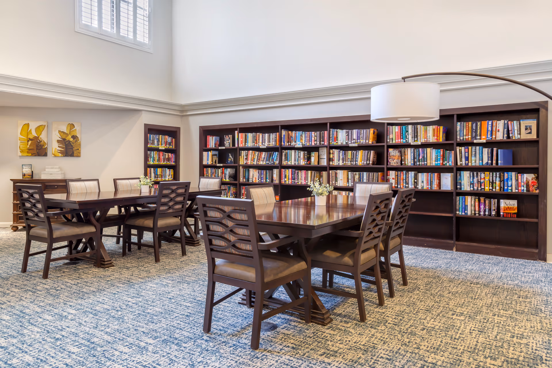 A bright and spacious library room with tall windows, featuring dark wooden bookshelves filled with books along the back wall. Two dark wooden tables with matching chairs are arranged in the center of the room, each table decorated with a small flower arrangement. The room has light-colored walls and a patterned carpet floor.