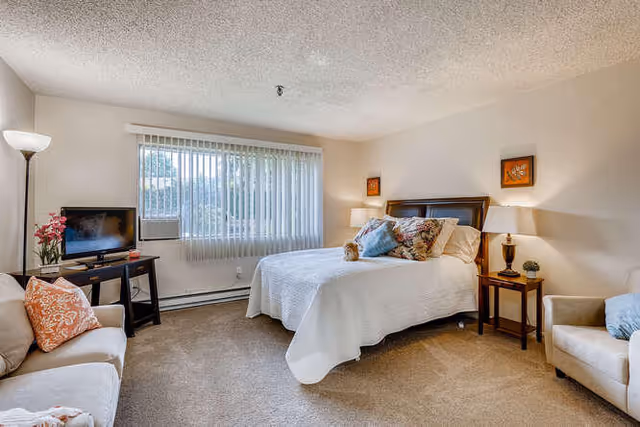 A cozy bedroom in Kamlu Retirement Inn-Vancouver featuring a neatly made bed with multiple pillows, two armchairs with cushions, a small TV on a stand, a floor lamp, and a window with vertical blinds letting in natural light.