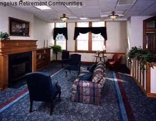 A living room area with patterned carpet, a floral sofa, two blue armchairs, a wooden fireplace with a framed picture above it, and windows with dark valances. There are plants and wooden furniture along the walls.