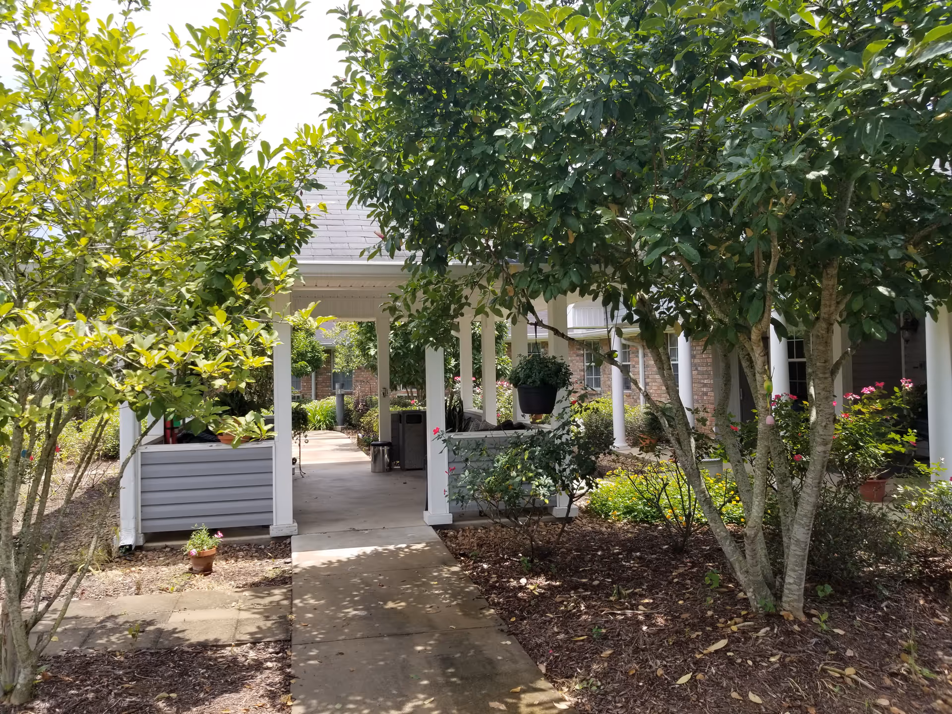 Covered walkway with white columns leading to a residential building entrance surrounded by trees and landscaped gardens.