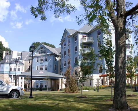 Exterior view of a multi-story senior living facility building with balconies, surrounded by trees and a grassy lawn under a partly cloudy sky. A white vehicle is parked near a covered entrance, and there are lampposts and landscaping around the building.