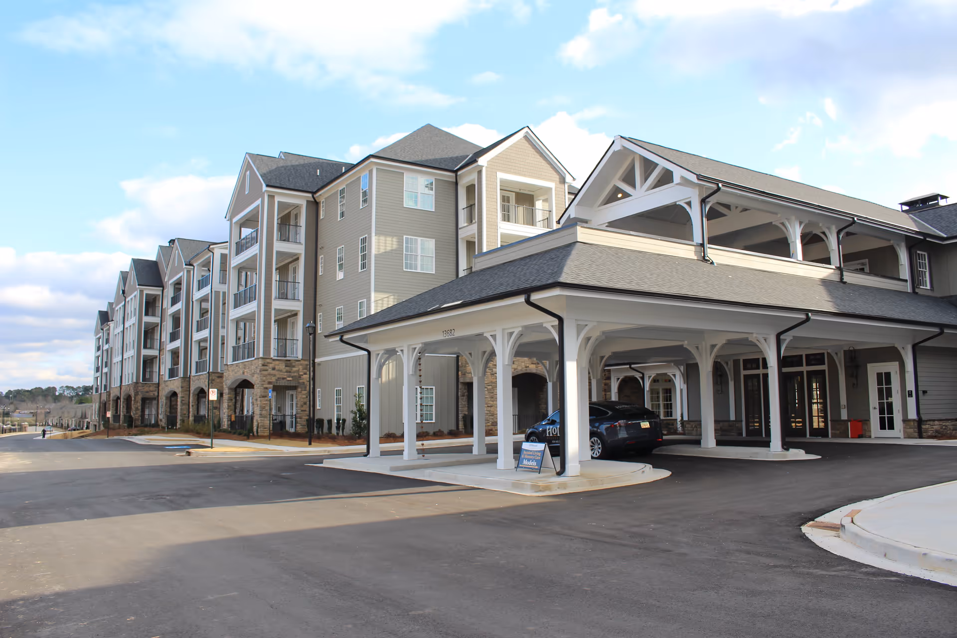 Exterior view of a large senior living facility building with multiple floors, balconies, and a covered entrance area. The building has beige siding with stone accents on the lower level, and a car is parked under the covered entrance. The sky is partly cloudy.